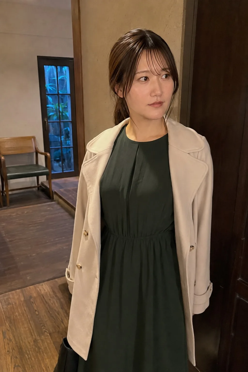 室内の入口で濃い緑のワンピースとベージュのジャケットを着て横を見る人物
