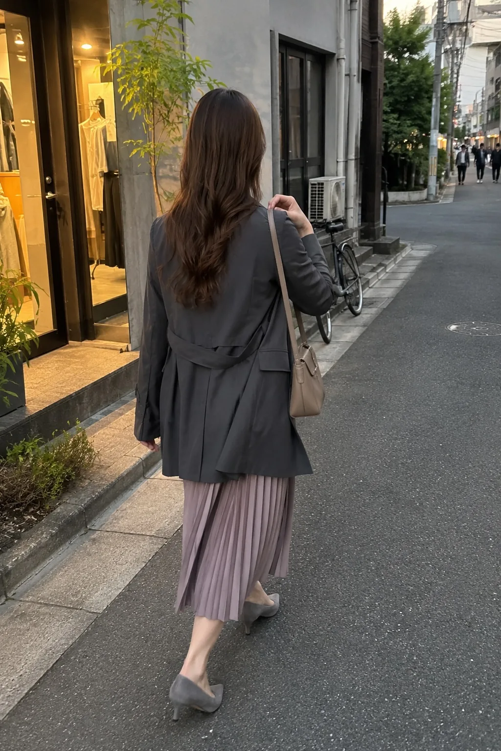店舗の並ぶ街路で濃いグレーのコートとプリーツスカートを着て歩く人物の後ろ姿