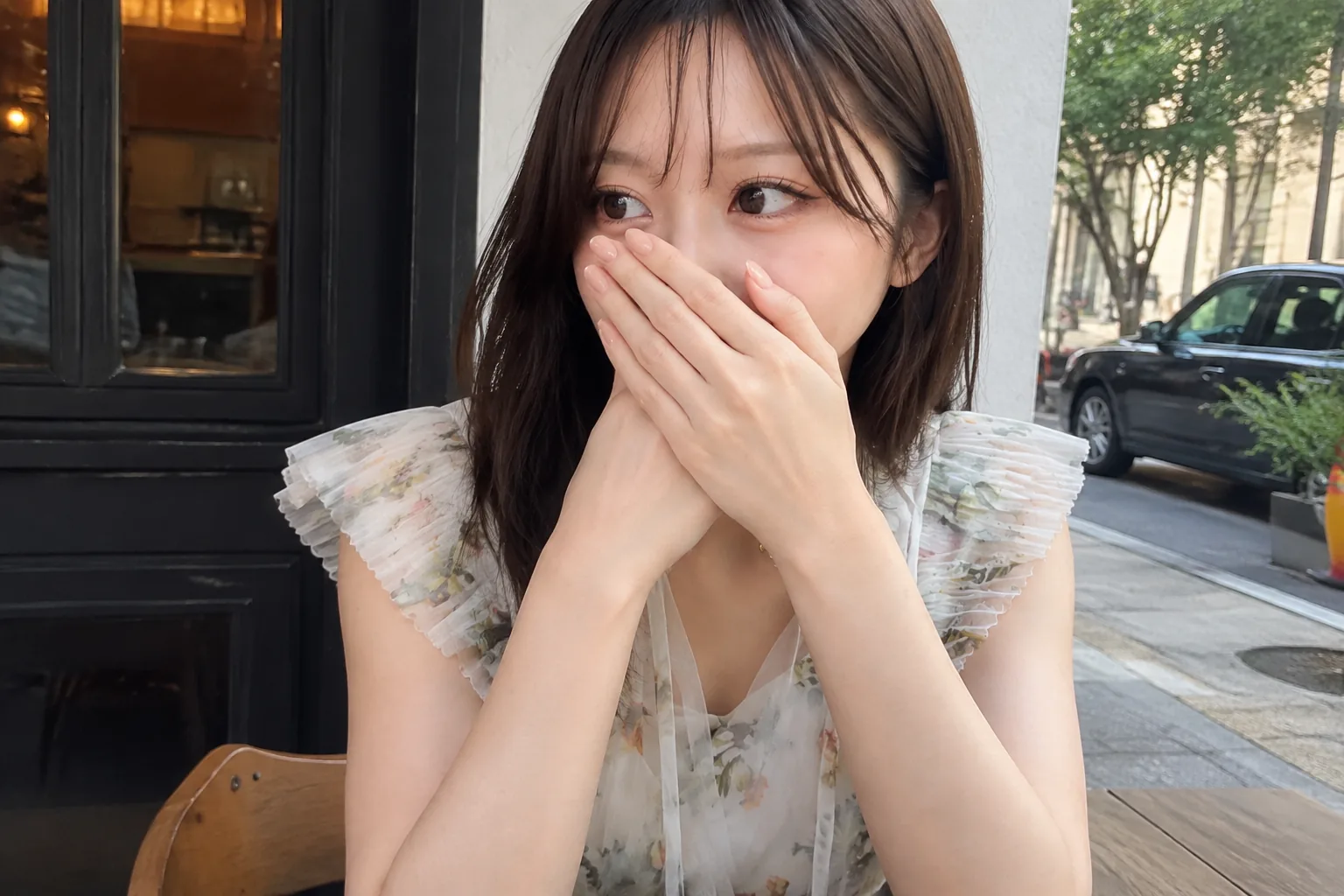 カフェのテラス席で花柄のワンピースを着て、重ねた両手を口元に当てて横を向く人物
