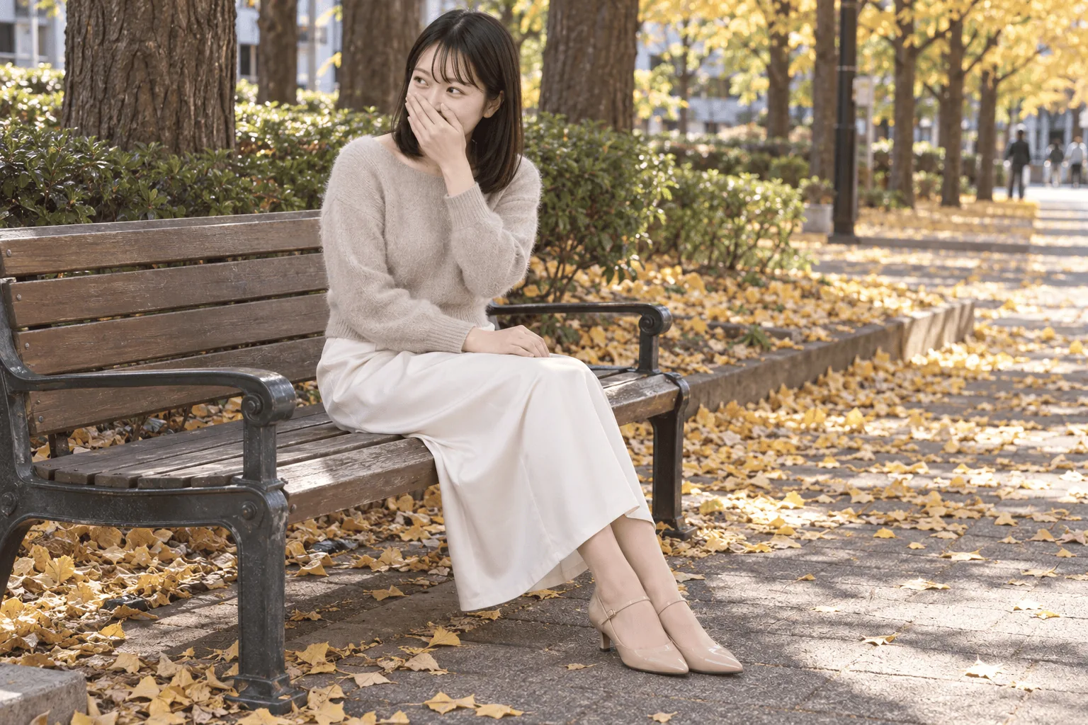落ち葉の積もる並木道のベンチに脚を揃えて座り、片手で鼻と口元を覆って画面左を見て笑う女性。もう片方の手は膝の上。横構図。